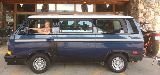 A blue vintage minivan parked outdoors in front of a stone building, with a woman leaning out of the driver's side window. A motorcycle is partially visible on the right.