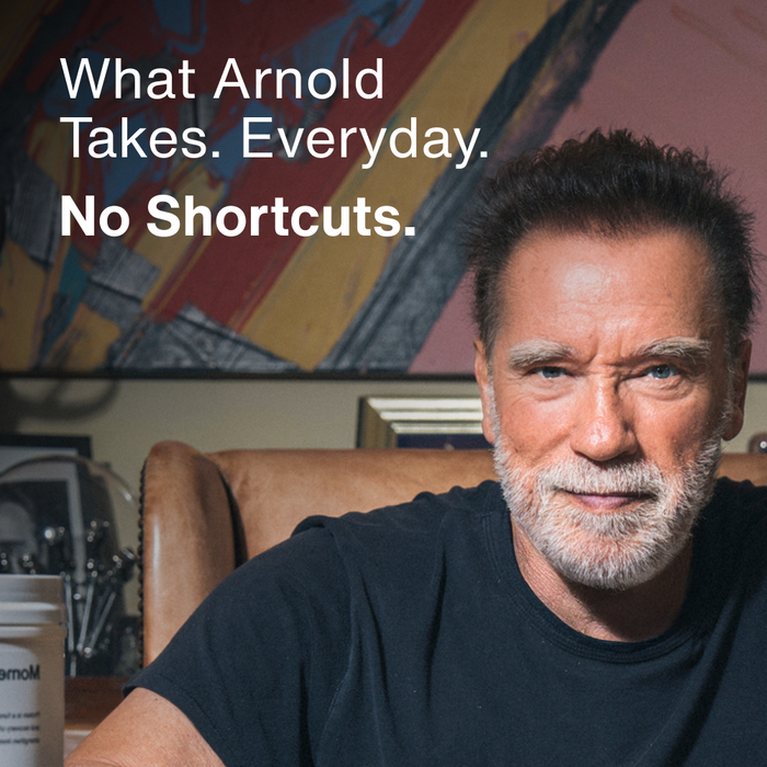 Arnold Schwarzenegger seated indoors with a Momentous product visible beside him. Text reads: What Arnold Takes. Everyday. No Shortcuts.