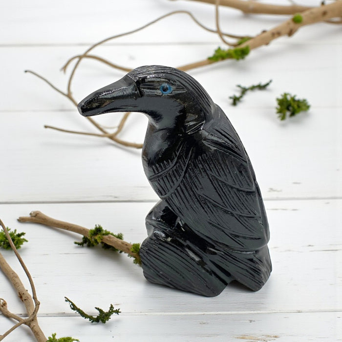 Black stone carving of a raven on a white wooden surface with branches and greenery.