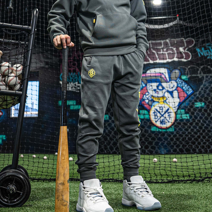 Person in gray tracksuit holding a baseball bat on a sports field with a colorful mural in the background.