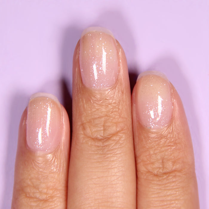 A close-up of fingers adorned with LONDONTOWN's Diamond Shine Top Coat, showcasing natural, glossy nails with a delicate diamond-dusted glittery finish against a light purple background.