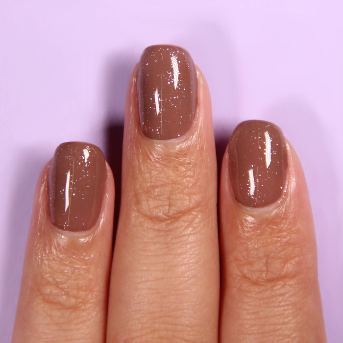 Fingers with short, neatly manicured nails painted in a glossy, light brown color and finished with LONDONTOWN's Diamond Shine Top Coat, featuring subtle diamond-dusted glitter, shown against a light purple background.