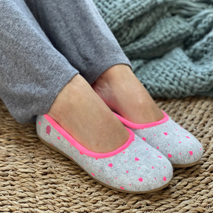 A pair of gray slippers with pink piping and heart shapes on the uppers, worn by a person sitting with their legs visible, wearing gray pants and a green sweater. Cosy comfortable Spanish slippers/house shoe.