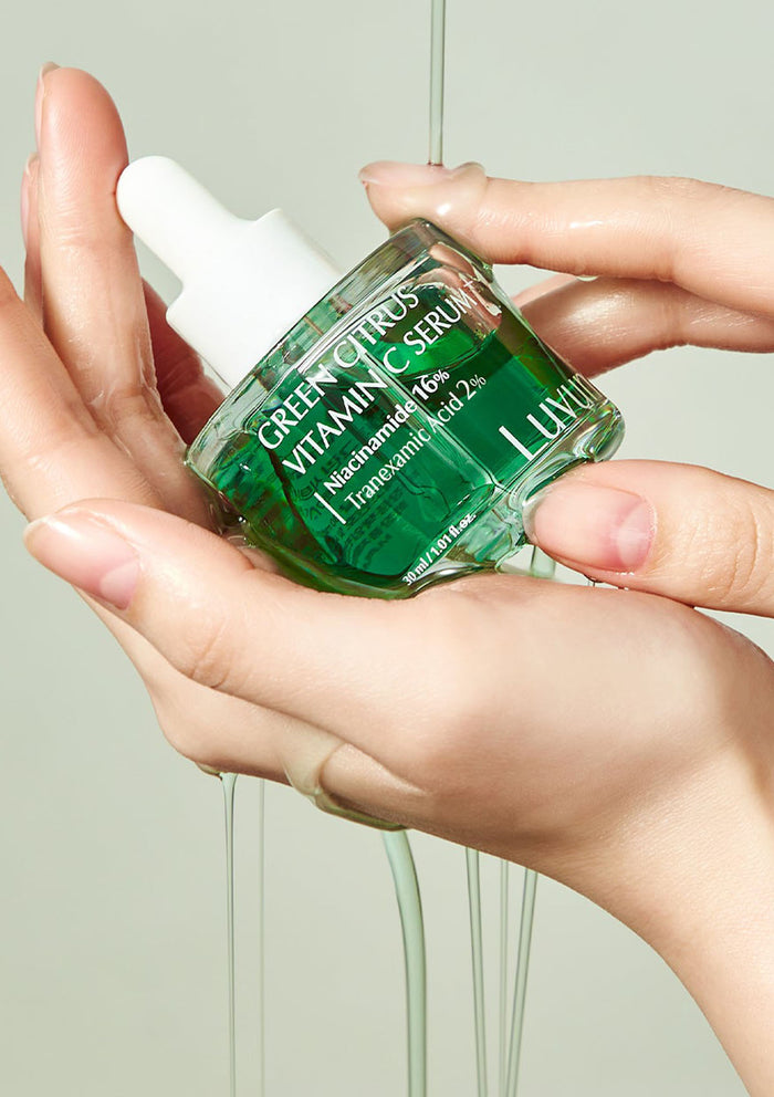 Hand holding a green skincare serum bottle labeled 'Green Citrus Vitamin C Serum' with a neutral background.