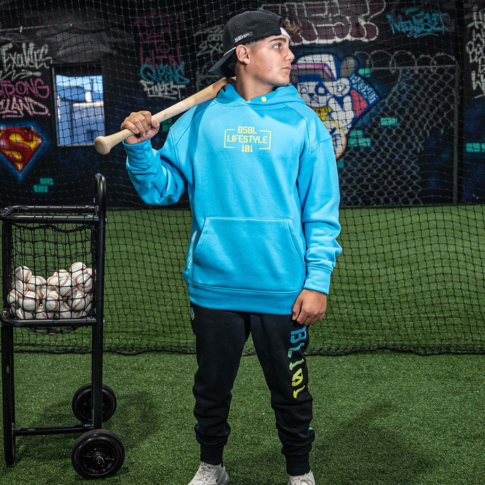 Person wearing a blue hoodie and black pants with 'BSBL LIFESTYLE 101' branding, holding a baseball bat in an indoor sports facility.