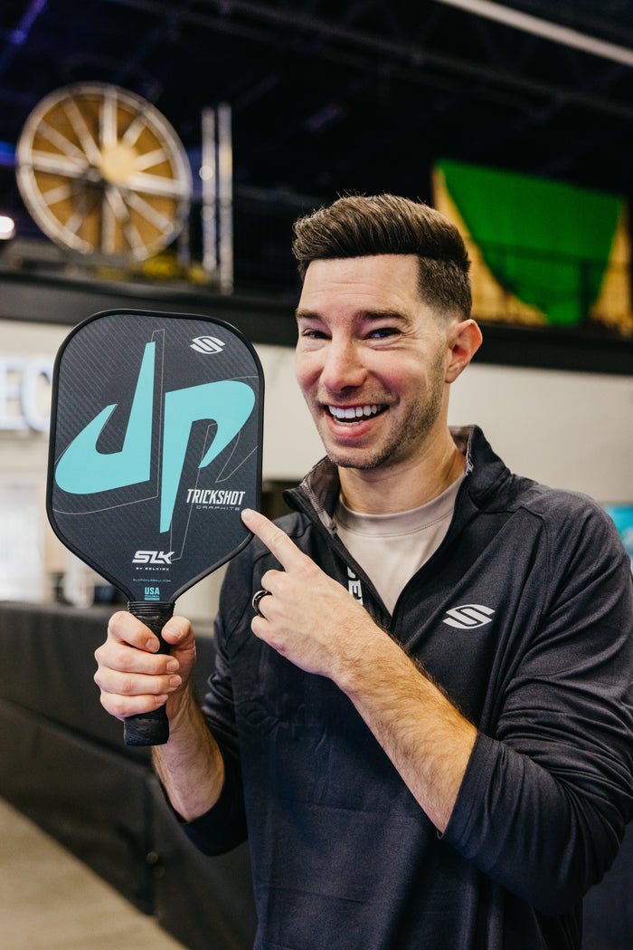 Smiling man pointing to a pickleball paddle labeled JP; paddle text reads TRICKSHOT SLK USA, jacket shows S logo.