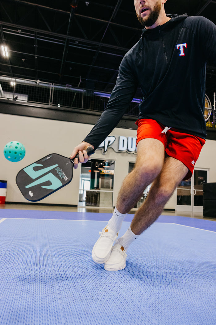 Player lunging to hit a pickleball with a paddle marked JP, wearing a black top with a T emblem.