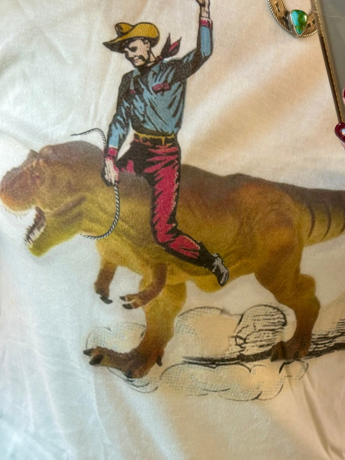 Light blue T-shirt close-up showing cowboy dinosaur riding graphic with boots and tail